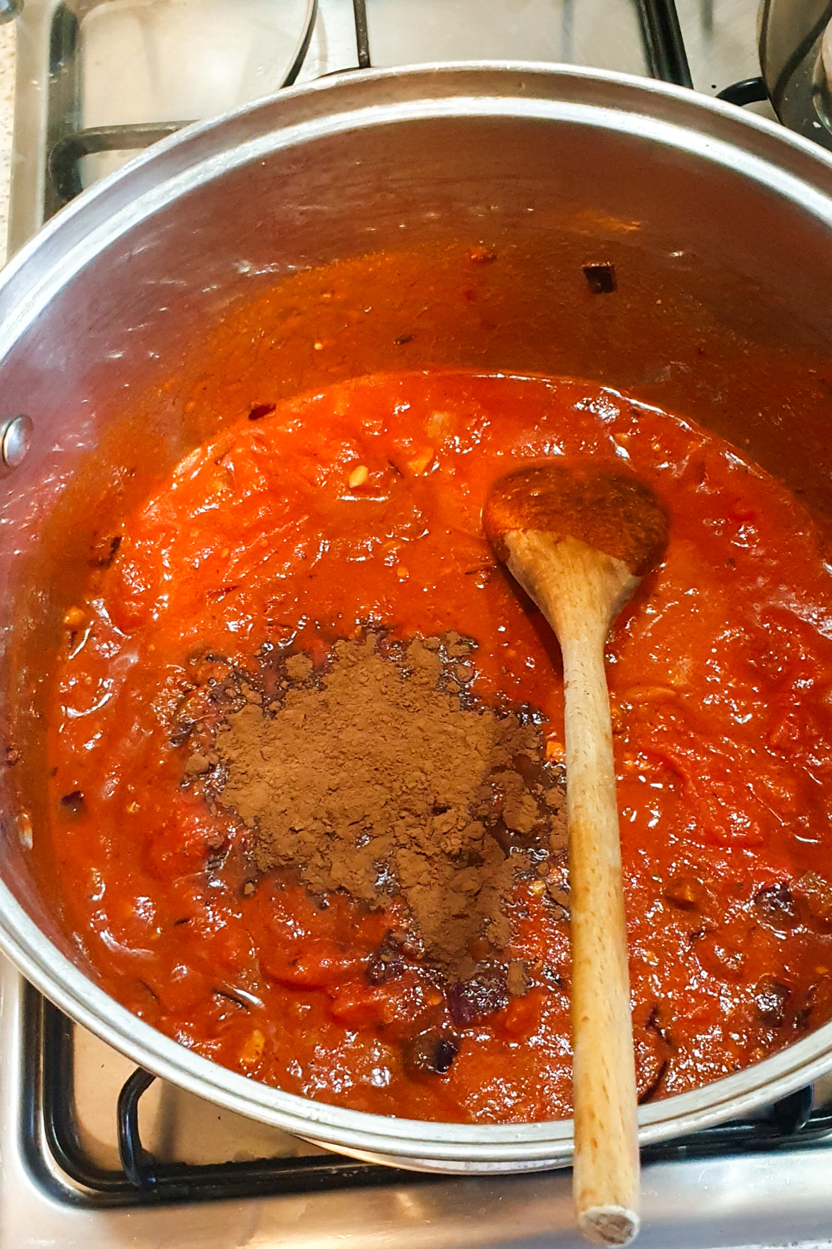 A pot of vegan chili simmers on the stovetop as a pile of cocoa powder is stirred in with a wooden spoon, creating a rich, high-protein tomato-based sauce.