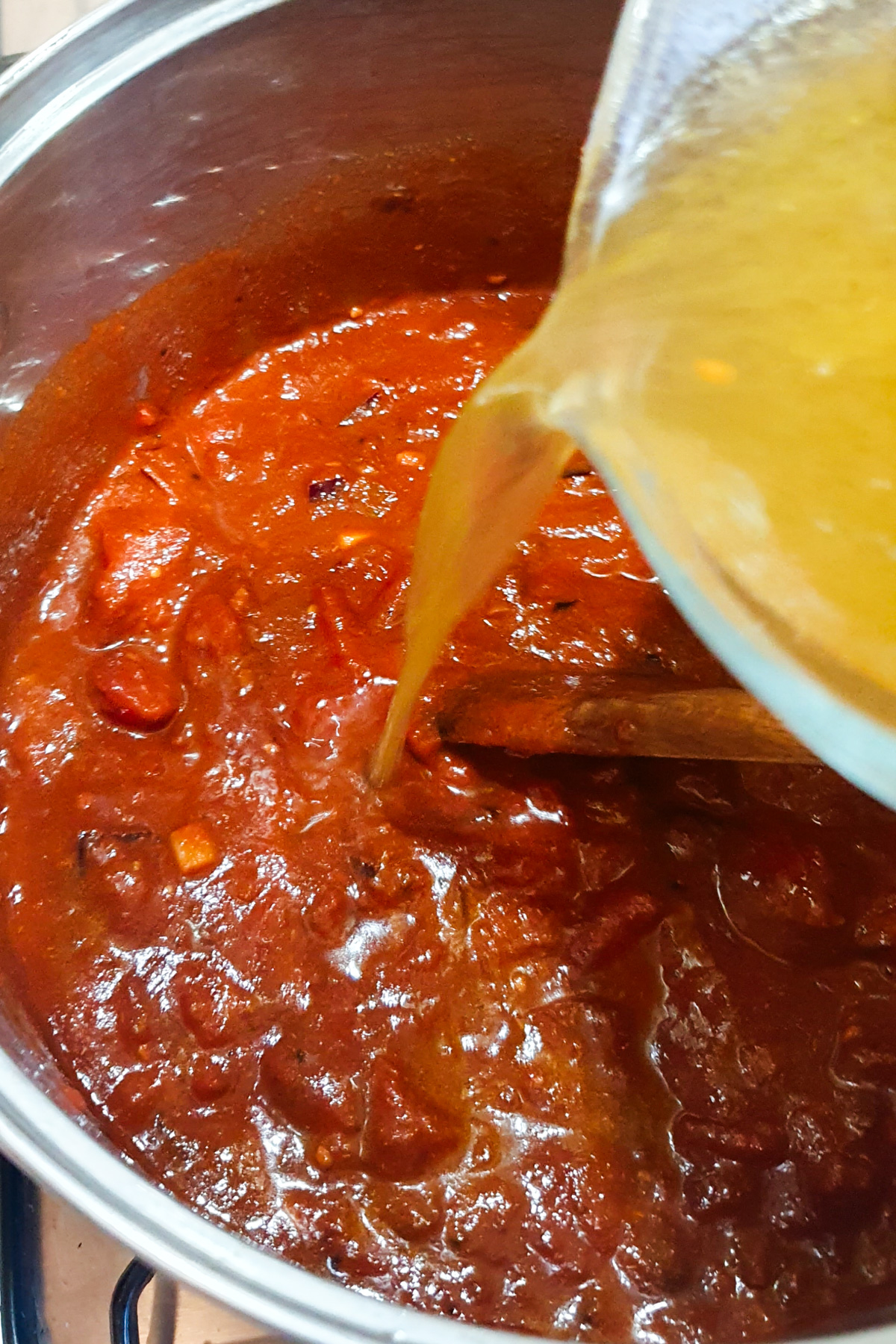 A pot of thick red vegan chili sauce is being stirred with a wooden spoon as yellow broth is poured in from a glass container.