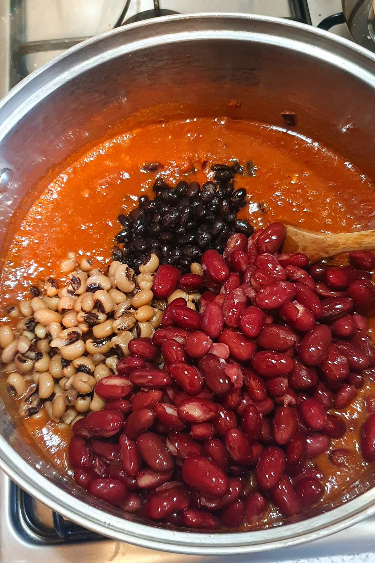 A pot of vegan chili simmers on the stovetop, packed with kidney beans, black-eyed peas, and black beans for a high protein meal, as they're stirred in with a wooden spoon.
