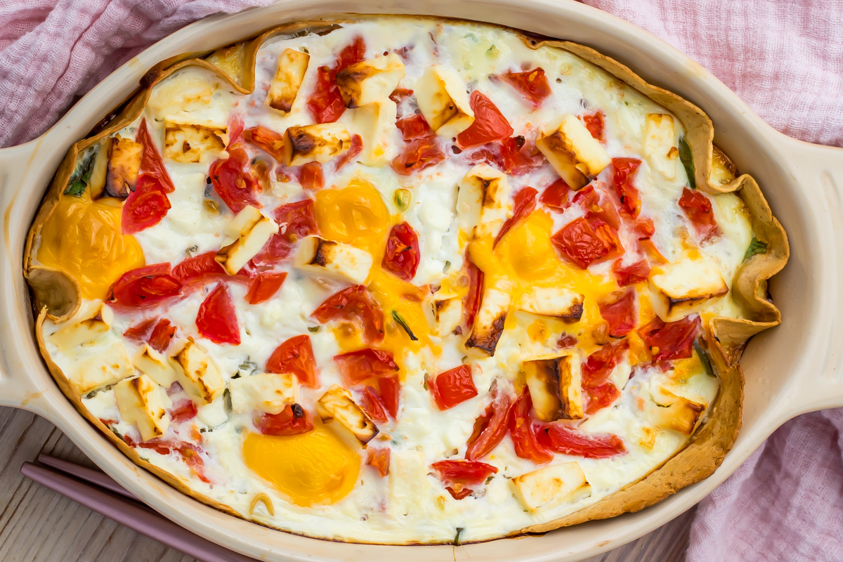 Egg Tortilla Bake: Baked dish with visible eggs, chopped tomatoes, and cubed cheese on a light crust, served in an oval ceramic baking dish on a pink cloth.
