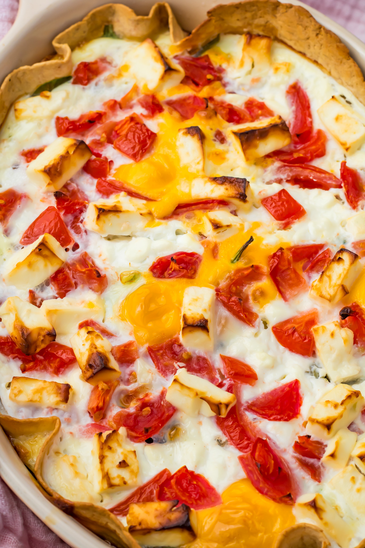 A close-up of an egg tortilla bake topped with melted cheese, tomatoes, and pieces of yellow egg yolk in a white dish.