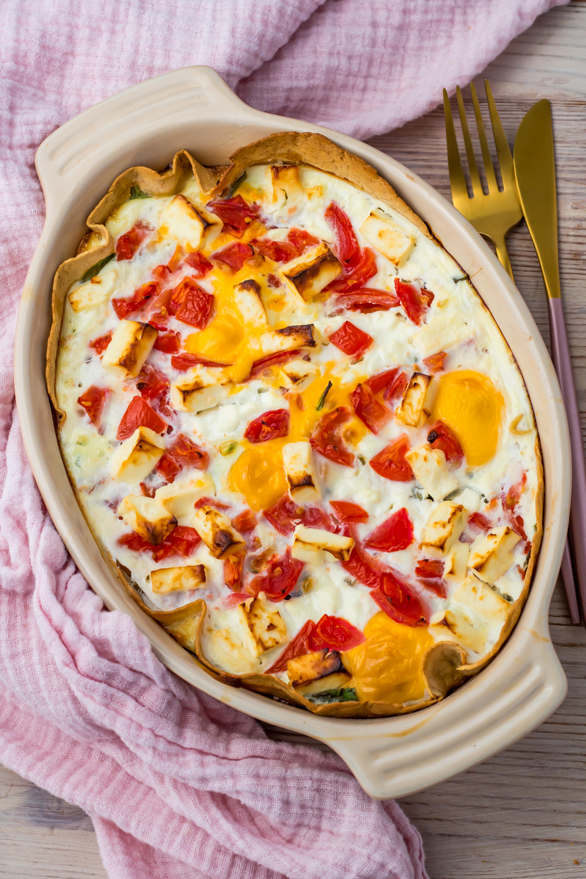 Egg Tortilla Bake casserole with tomatoes and cheese in a beige oval dish, placed on a pink cloth with gold cutlery beside it.