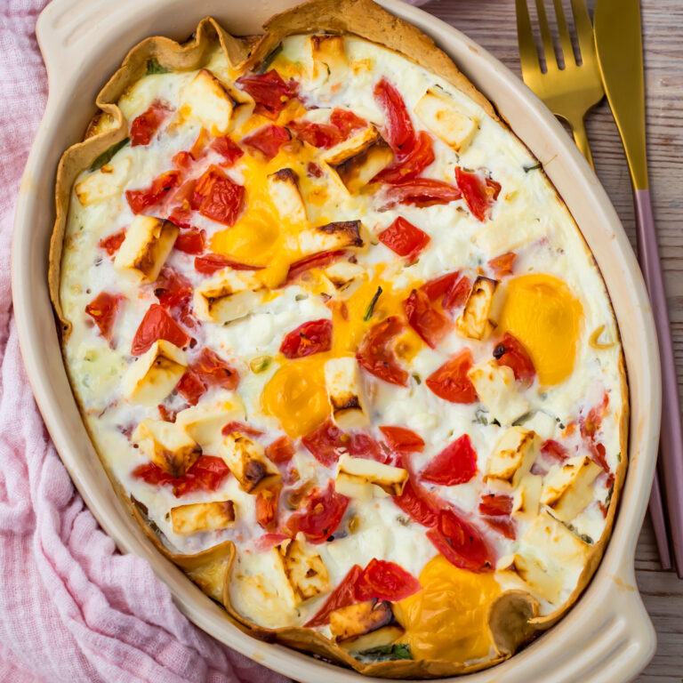 An Egg Tortilla Bake casserole with baked eggs, tomatoes, yellow bell peppers, and cheese in a beige oval dish, placed on a pink cloth beside a gold fork and knife.