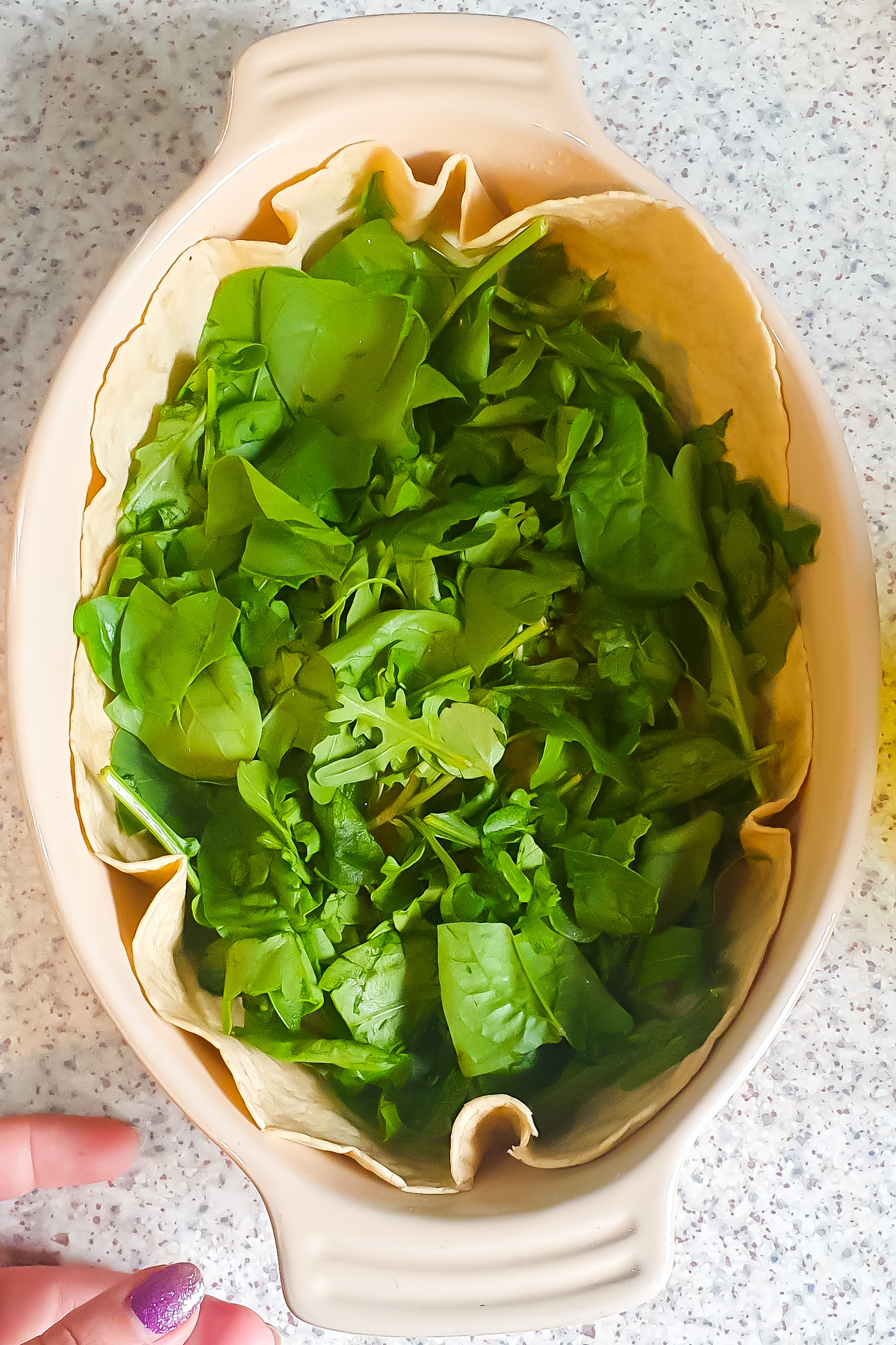 A baking dish lined with a tortilla and fresh spinach leaves, ready for an Egg Tortilla Bake, sits on a speckled countertop. A hand with purple-painted nails is visible at the bottom left.