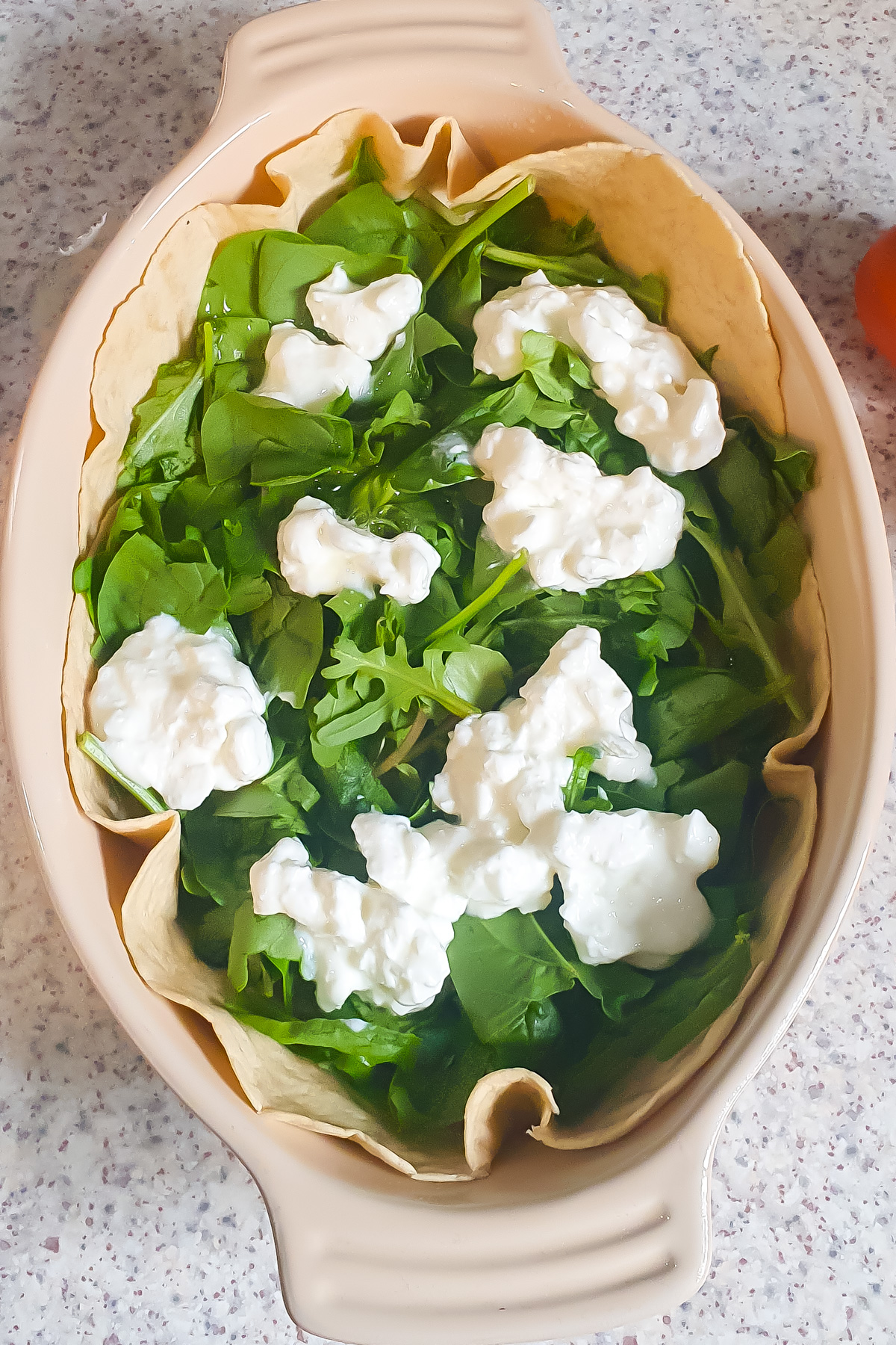 A baking dish lined with a tortilla, filled with fresh spinach leaves and topped with cottage cheese, creates a delicious Egg Tortilla Bake sitting on a speckled countertop.