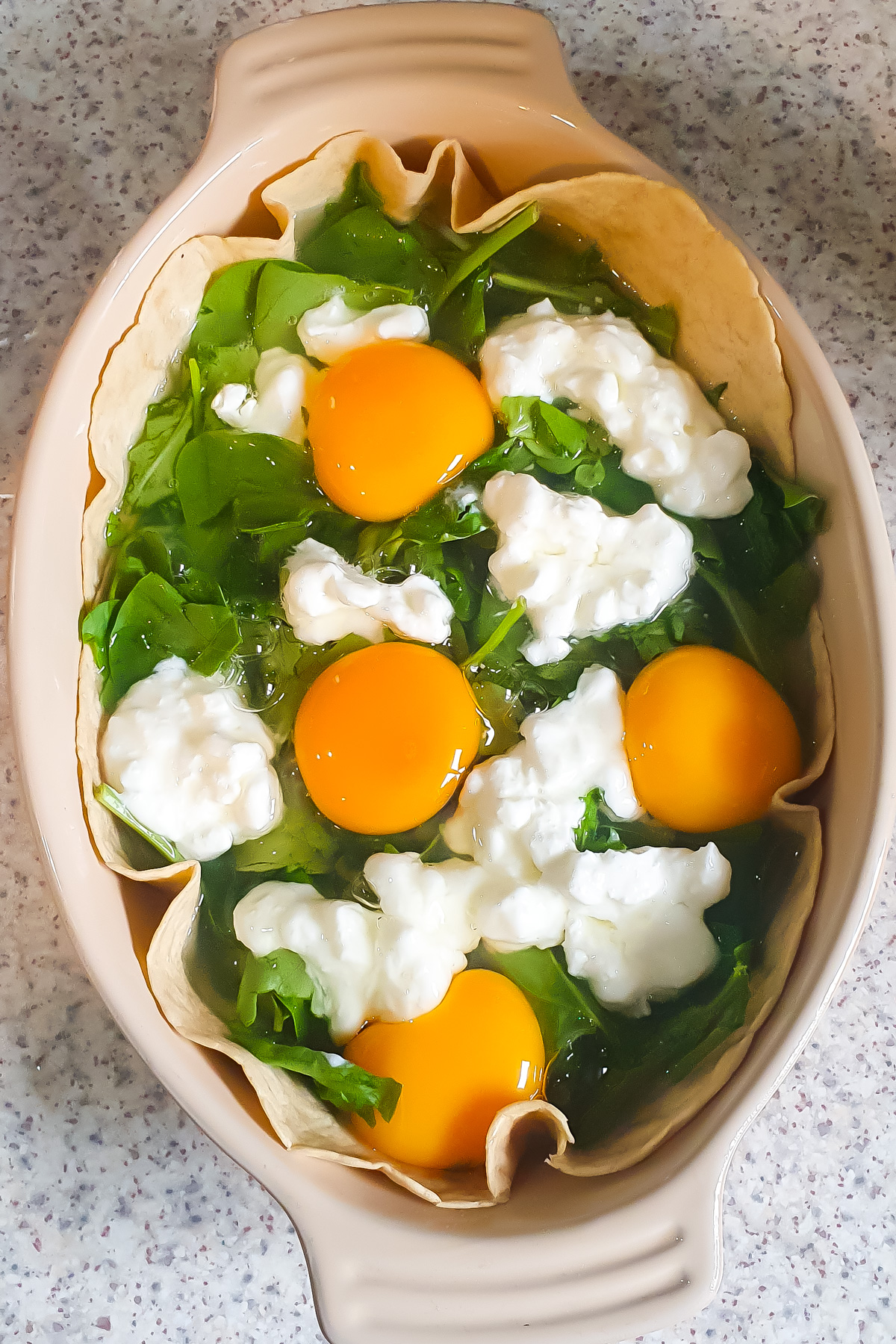A baking dish lined with tortilla, filled with fresh spinach leaves, raw eggs, and dollops of cottage cheese—this Egg Tortilla Bake sits ready on a speckled countertop.