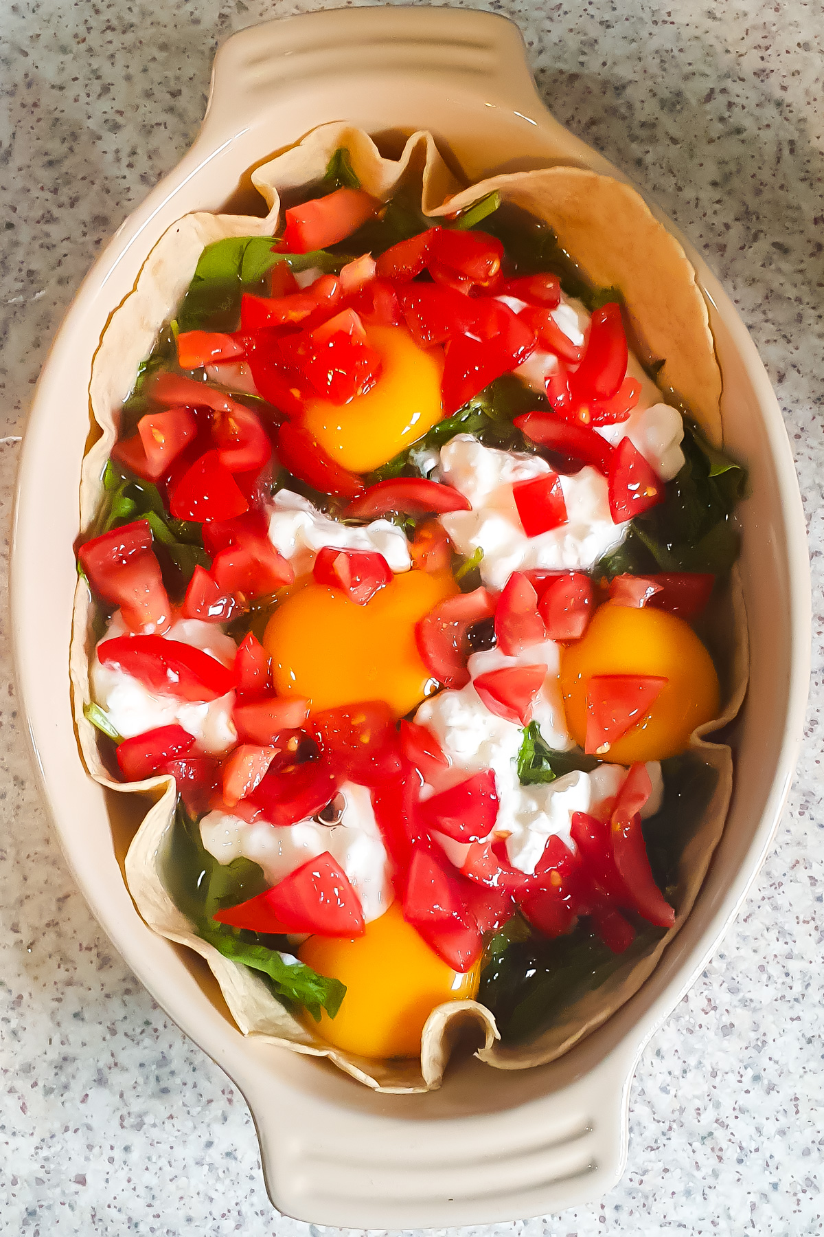 An oval baking dish containing ingredients for Egg Tortilla Bake—tortillas, raw eggs, chopped spinach, tomatoes, and cottage cheese—rests on a speckled countertop.