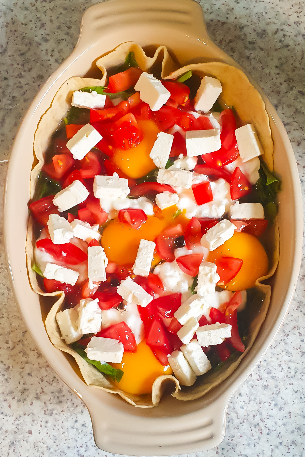 An oval baking dish with an unbaked Egg Tortilla Bake—eggs, diced tomatoes, spinach, and rectangular pieces of white cheese on a tortilla crust—posed on a speckled countertop.