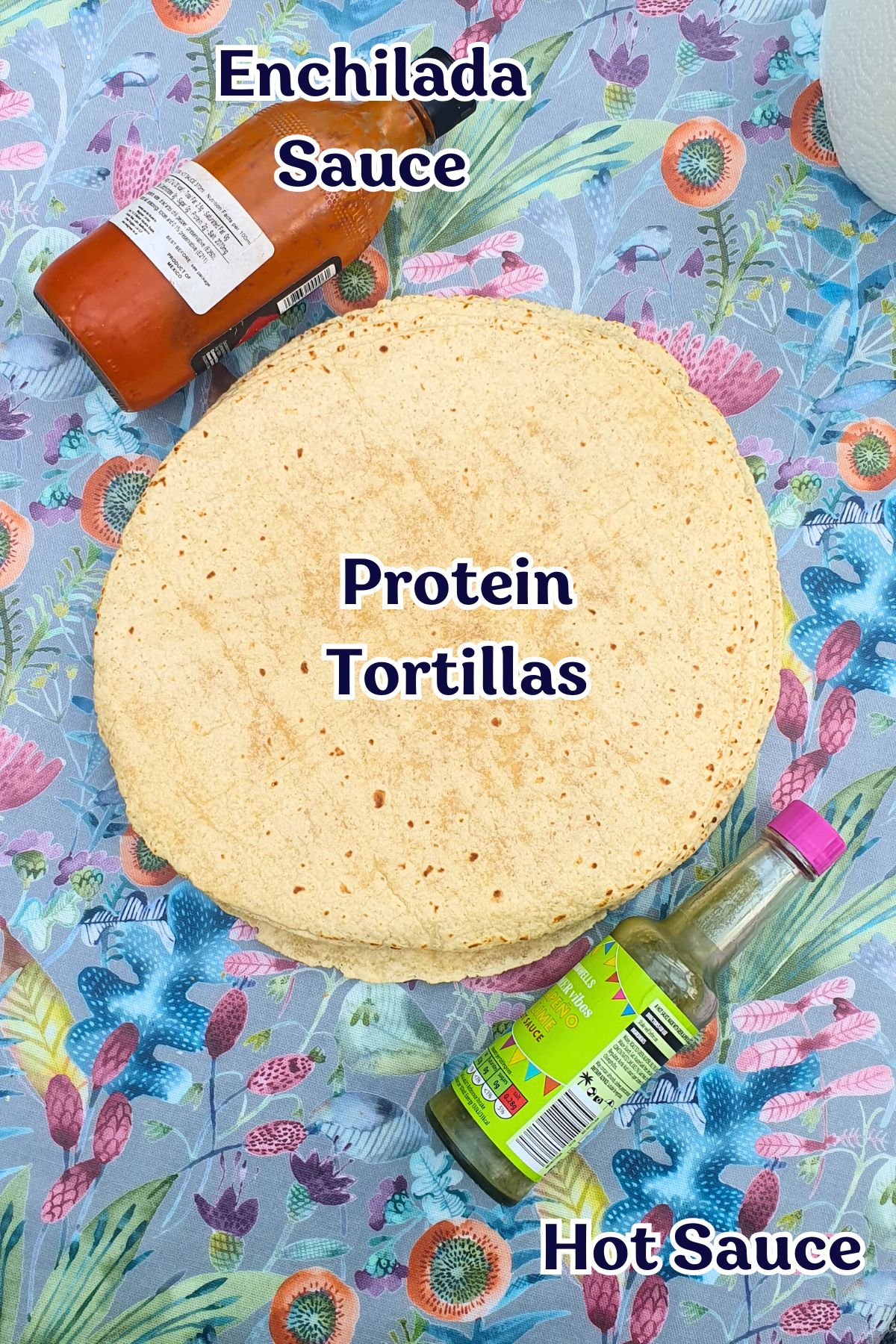 A stack of high protein tortillas on a floral tablecloth, with a bottle of enchilada sauce above and a bottle of hot sauce below, both labeled.