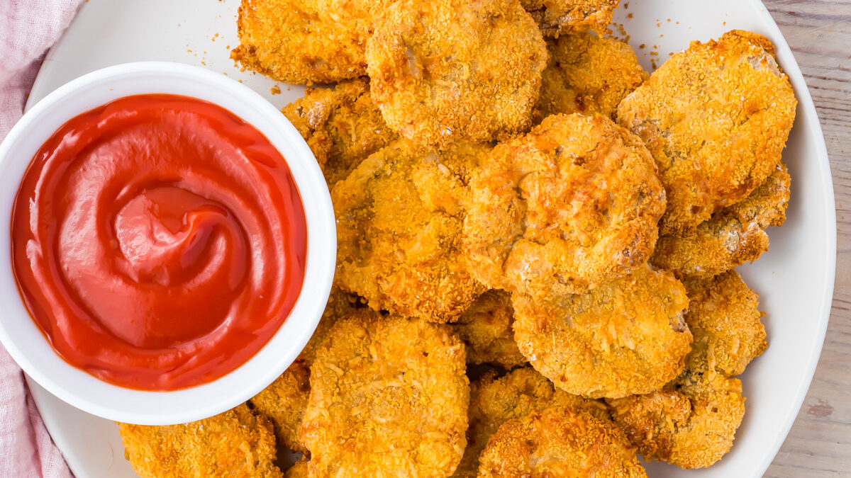 A white plate filled with crispy Air Fryer homemade chicken nuggets made from ground chicken sits next to a small bowl of red dipping sauce, placed on a light wooden surface.