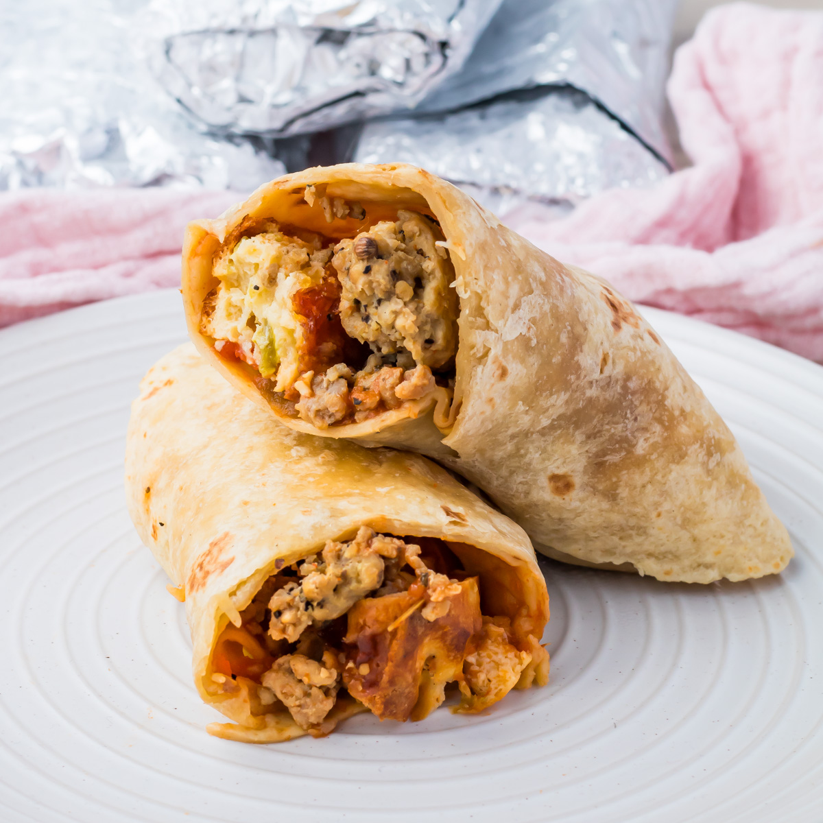 Two high protein breakfast burritos filled with scrambled eggs, ground meat, and vegetables are stacked on a white plate, with foil-wrapped burritos and a pink towel in the background.