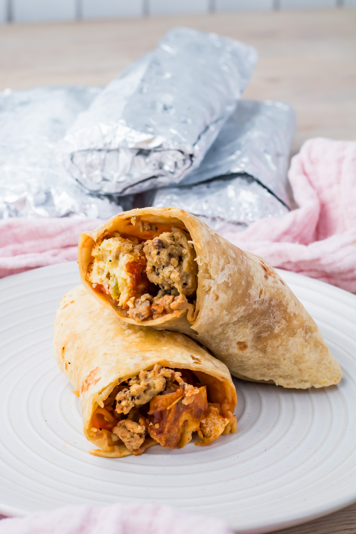 Two high protein breakfast burritos cut in half reveal scrambled eggs, sausage, and vegetables inside. They&rsquo;re stacked on a white plate, with more foil-wrapped burritos in the background&mdash;perfect for a quick breakfast recipe.