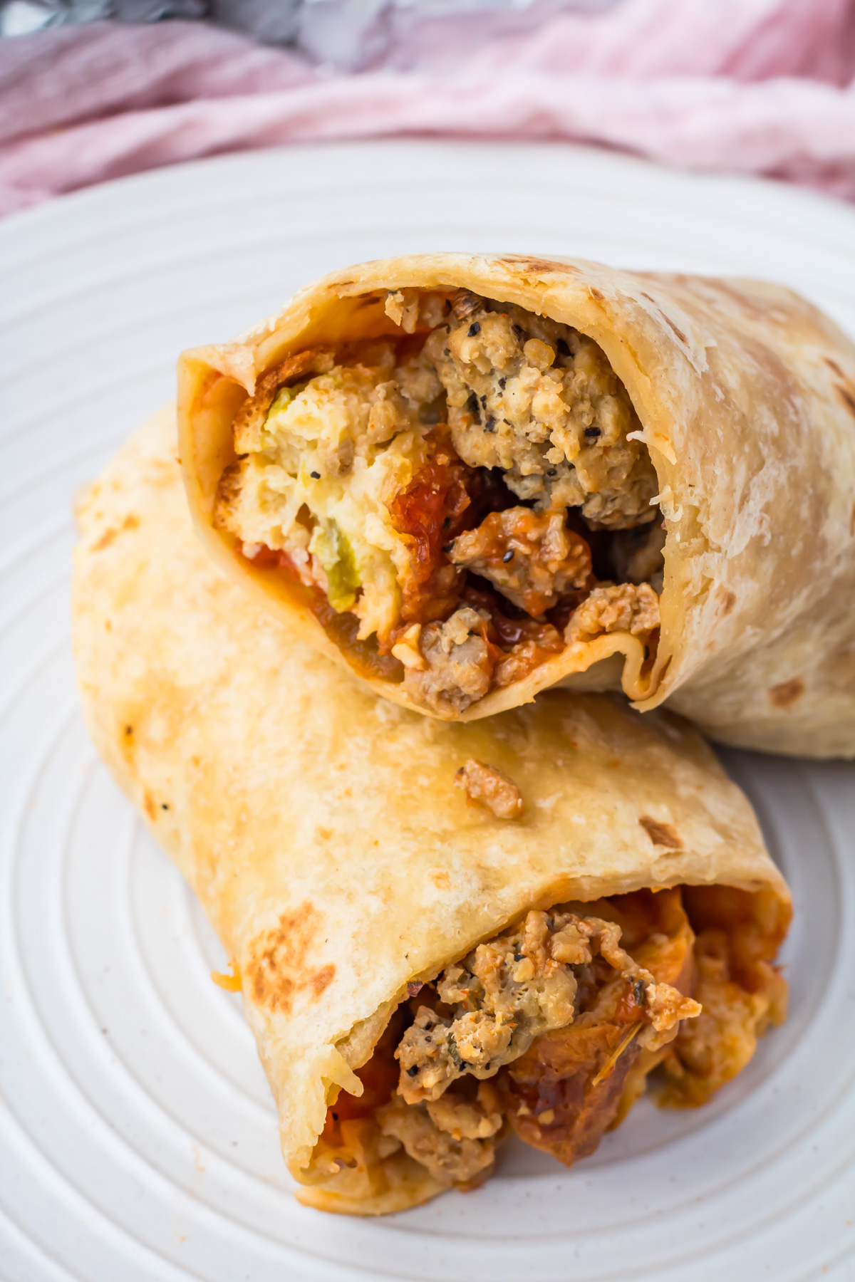 A high protein breakfast burrito cut in half reveals scrambled eggs, ground meat, cheese, and vegetables tucked inside a flour tortilla on a white plate&mdash;an easy recipe to start your morning right.