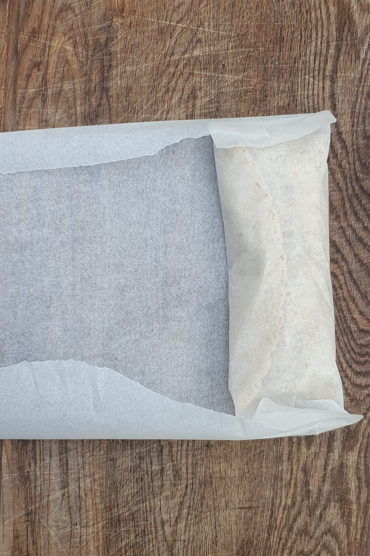 A rectangular piece of dough, resembling a high protein breakfast burrito, wrapped in parchment paper on a wooden surface, partially enclosed by the paper.