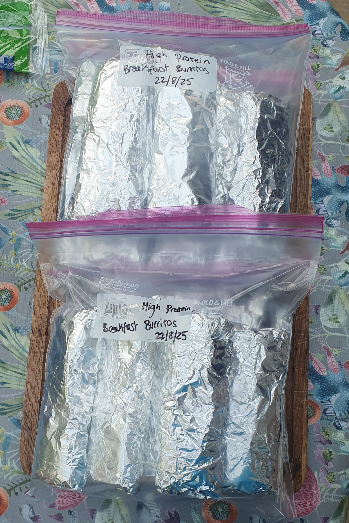 Two ziplock bags labeled "High Protein Breakfast Burritos 22/8/25," each filled with several foil-wrapped high protein breakfast burritos, are neatly placed on a wooden board.