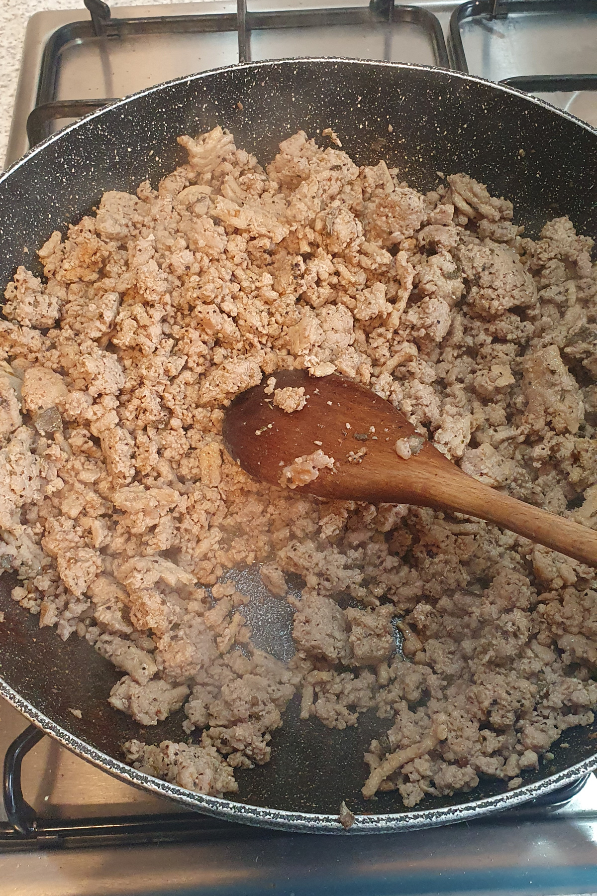 A frying pan with cooked ground meat for a high protein breakfast burrito is being stirred by a wooden spoon on a gas stove.