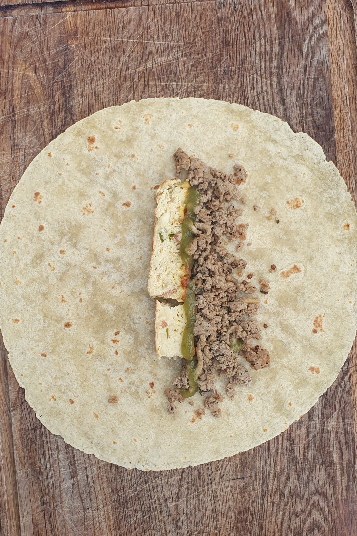 A high protein breakfast burrito rests on a wooden surface, filled with a strip of scrambled eggs, ground meat, and green salsa down the center.