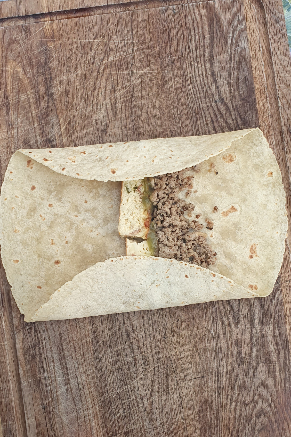 A high protein breakfast burrito sits on a wooden board, partially filled with ground meat, tofu, and vegetables, its sides folded in but not fully wrapped.