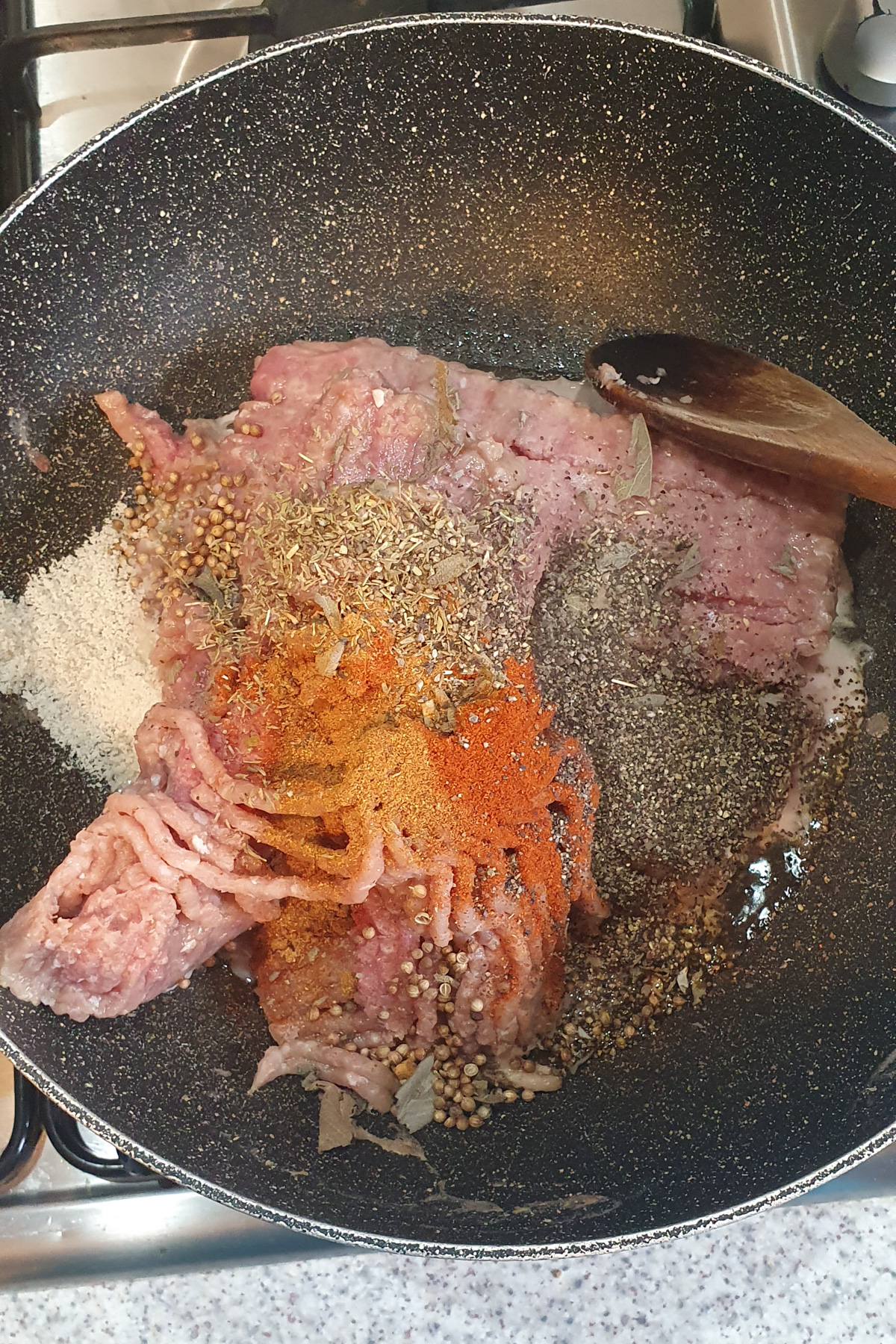 Raw ground meat in a frying pan with various spices, seasonings, and a wooden spoon on a stovetop.