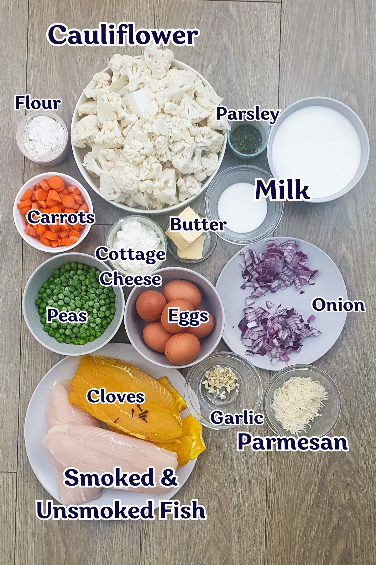 Various labeled ingredients for a fish pie are arranged on a table, including cauliflower, carrots, peas, eggs, onion, fish fillets, milk, cheese, butter, flour, garlic, parsley, and parmesan.