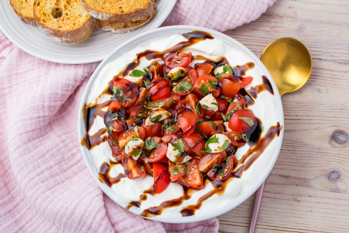 A bowl of whipped cottage cheese topped with chopped tomatoes, basil, and balsamic glaze, sits beside toasted bread slices on a wooden table with a pink cloth.