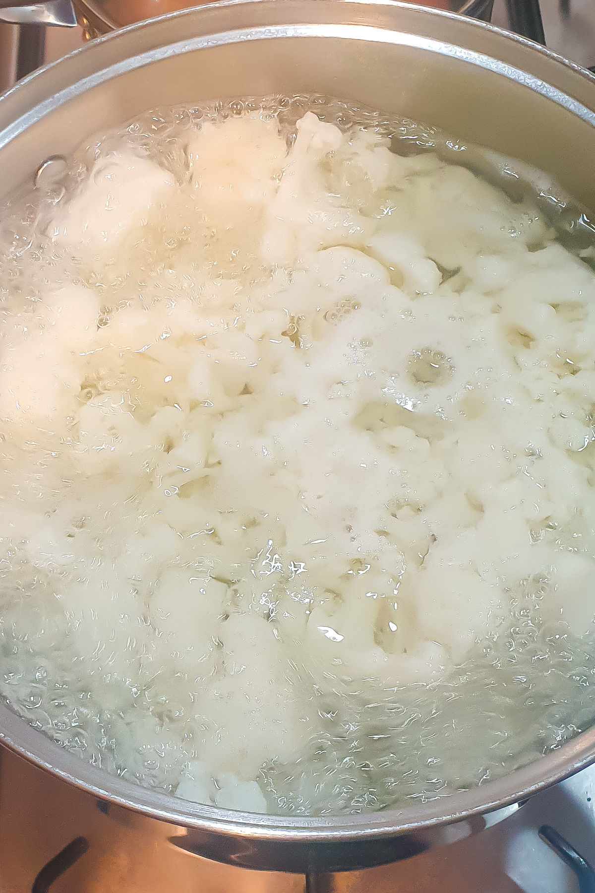 Cauliflower pieces are boiling in water inside a stainless steel pot on a stove, the first step in this classic recipe.