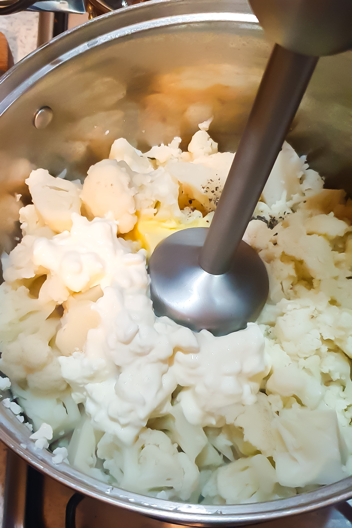 An immersion blender is blending cooked cauliflower, butter, and cottage cheese in a pot.