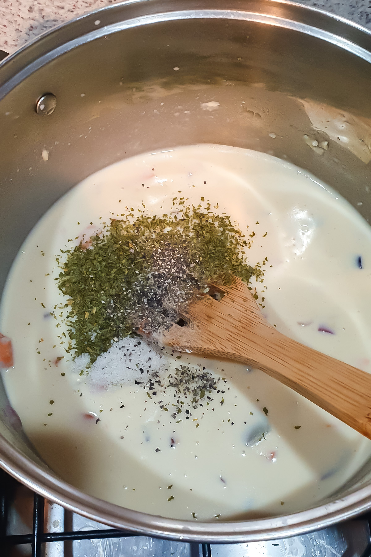 A pot of creamy white sauce being stirred with a wooden spoon, with dried herbs and seasonings sprinkled on top.