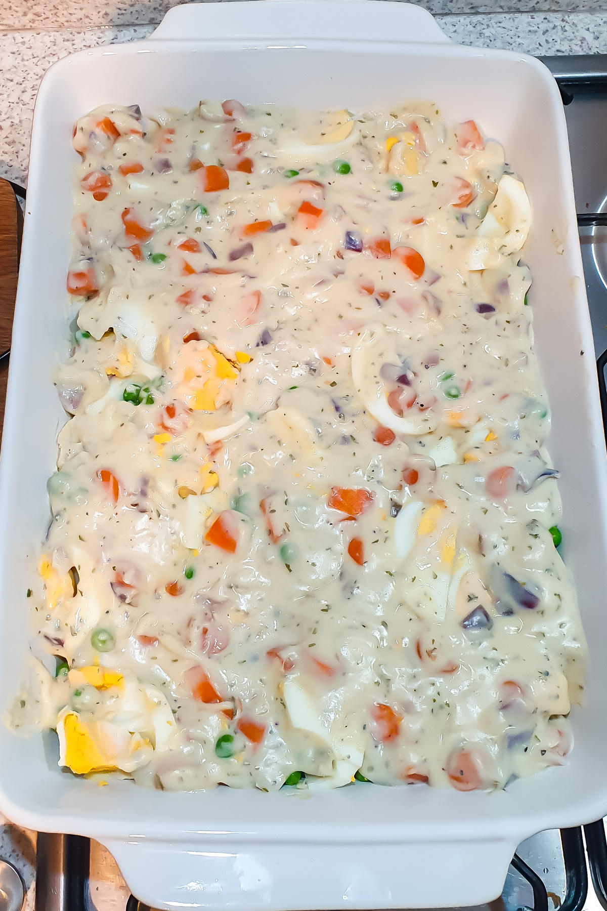 A white baking dish filled with an unbaked creamy mixture of vegetables, sliced eggs, and sauce.
