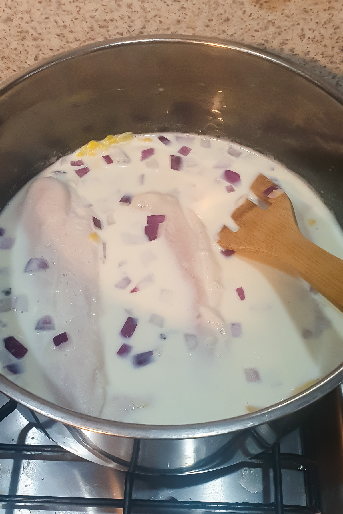 Raw fish fillets, chopped red onion, and milk in a pot on a stove with a wooden spatula.
