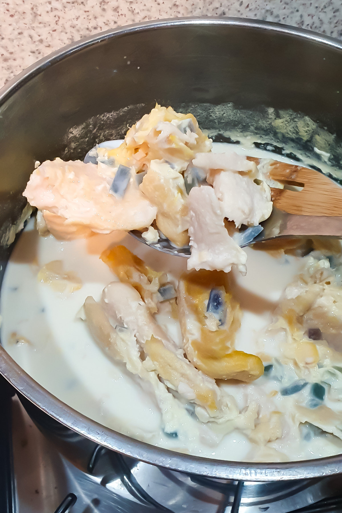 A pot with chunks of fish, vegetables, and milk being stirred with a wooden spoon on a stovetop.