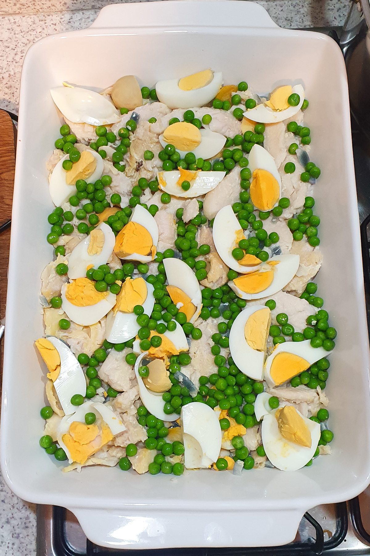 A white baking dish filled with cooked fish, hard-boiled egg wedges, and green peas in a single layer.