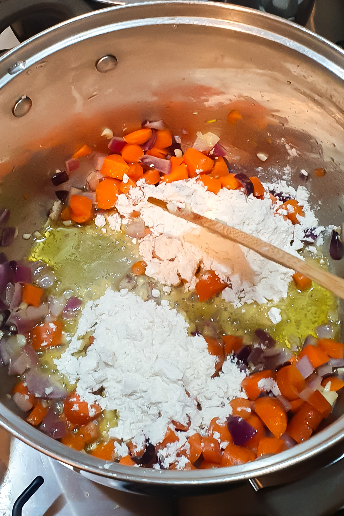 Chopped carrots and red onions are sautéing in a pot with oil, with flour sprinkled on top and a wooden spoon resting inside.