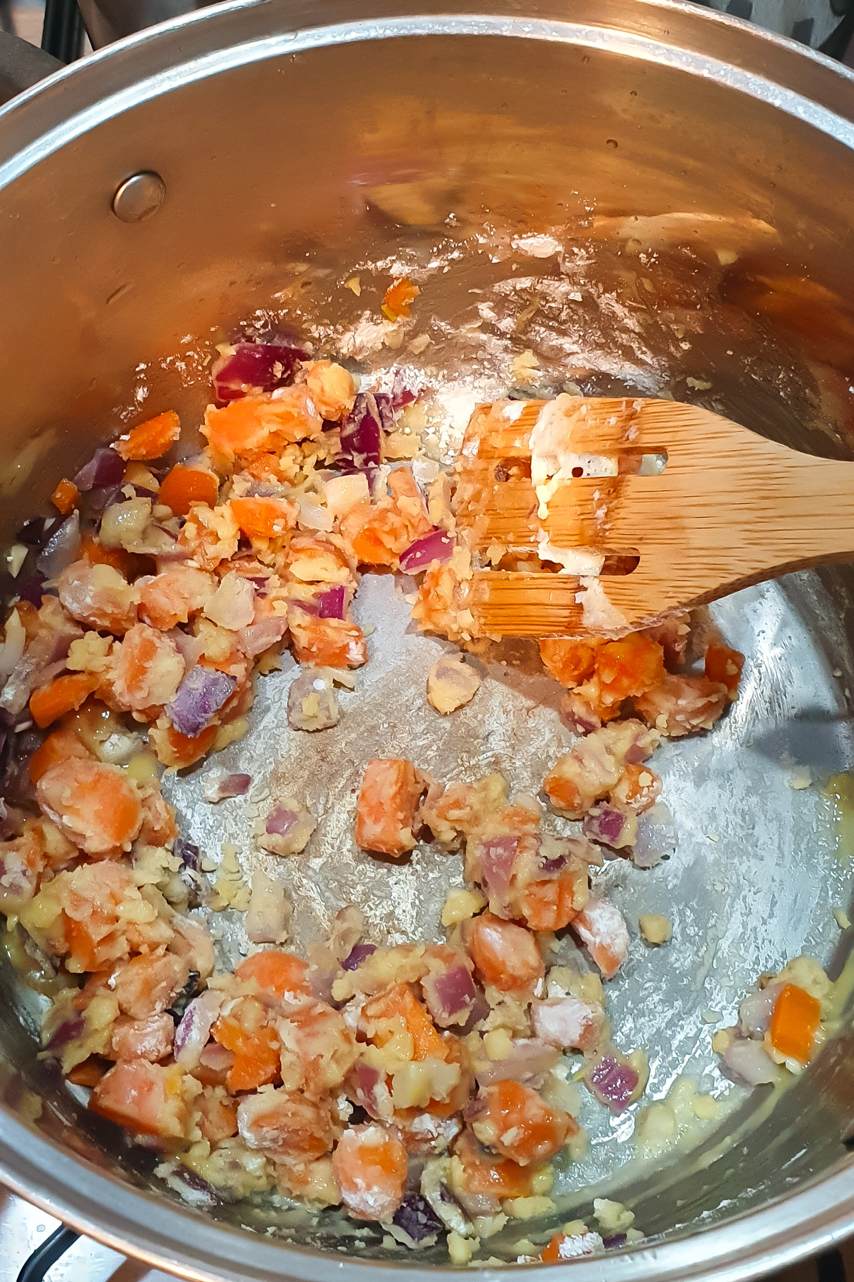 Chopped carrots, onions, and garlic sautéing in a pot with a wooden spatula, with flour mixed in.