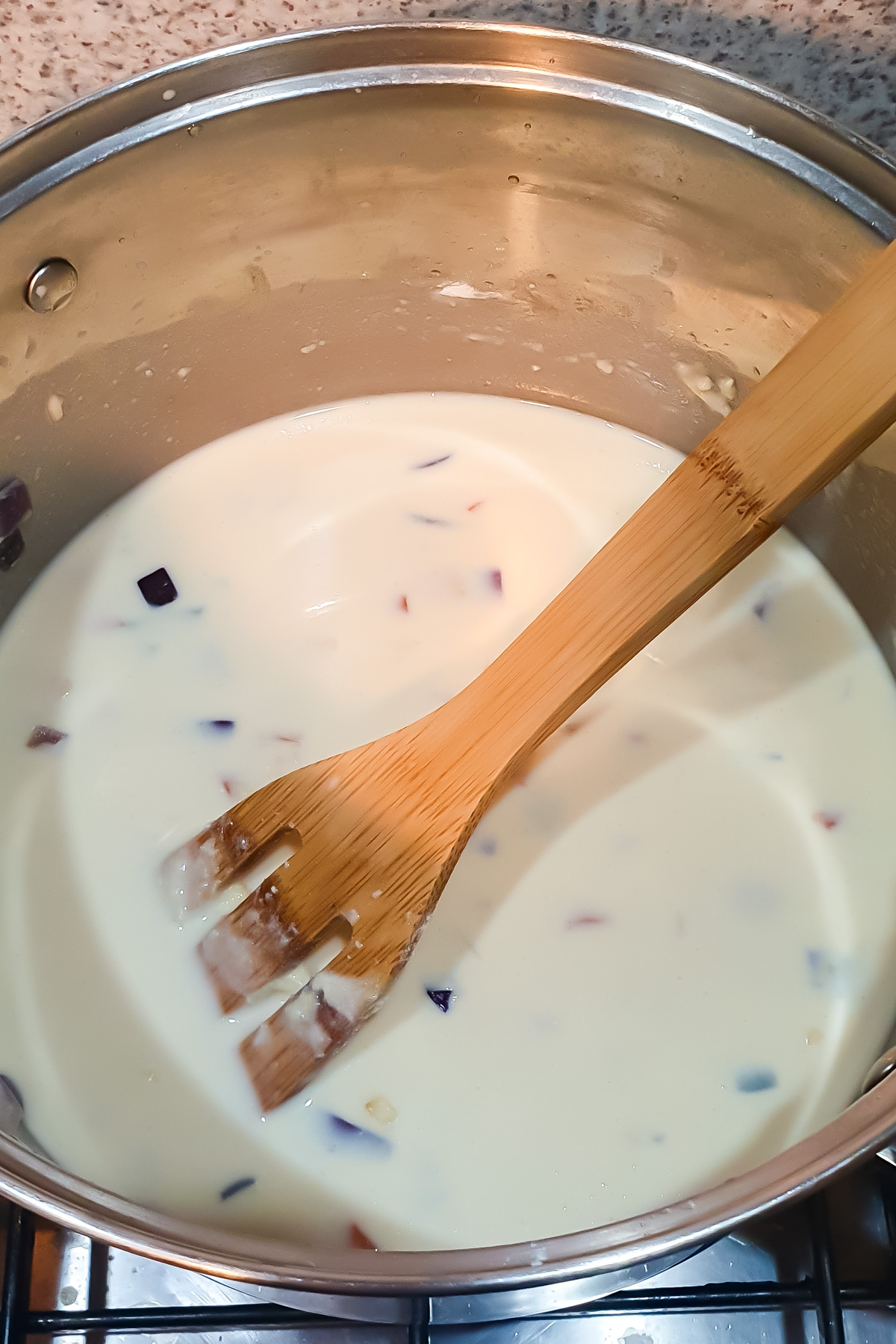 A wooden spoon stirs a creamy white sauce with small colorful vegetable pieces in a stainless steel pot on a stove.