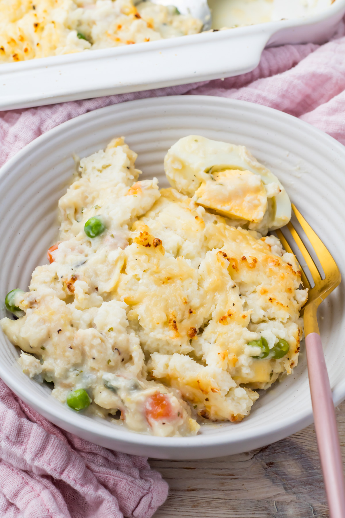 A bowl of high protein Fish Pie casserole with cauliflower, peas, carrots, and a creamy sauce. It is served with a fork resting inside the dish.