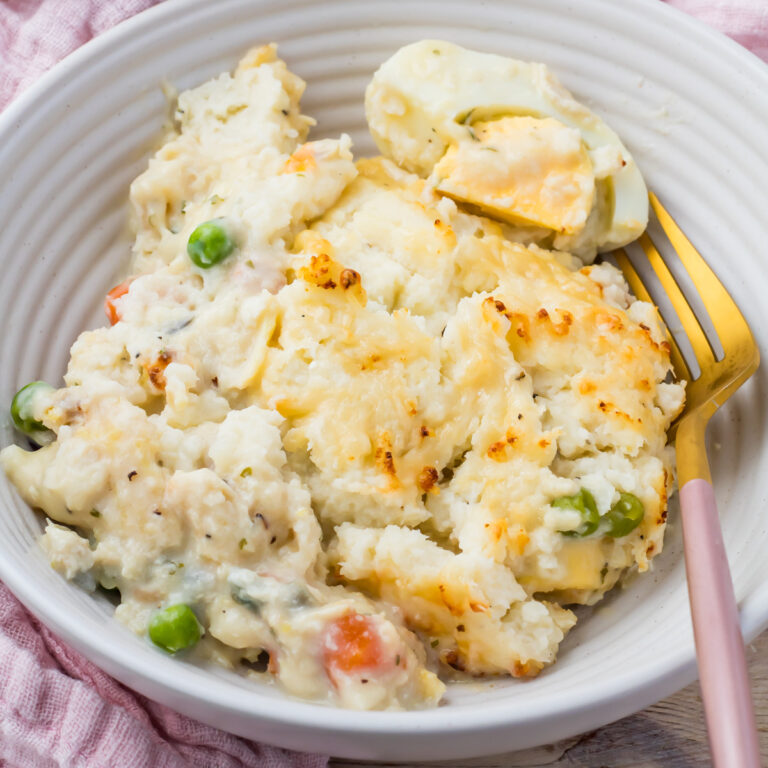 A serving of creamy, high protein fish pie with peas and carrots on a white plate, next to a gold fork.