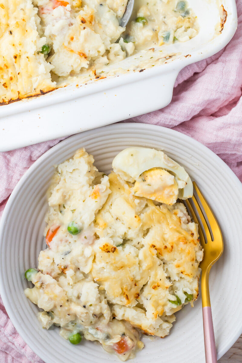 A serving of high protein fish pie with a mashed cauliflower topping on a plate sits next to a baking dish with the remaining casserole.