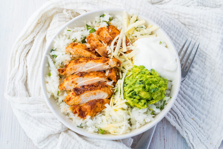 A white bowl with sliced grilled chicken, shredded cheese, guacamole, sour cream, and rice sits on a white cloth with a fork&mdash;perfect for those seeking high protein chicken recipes.
