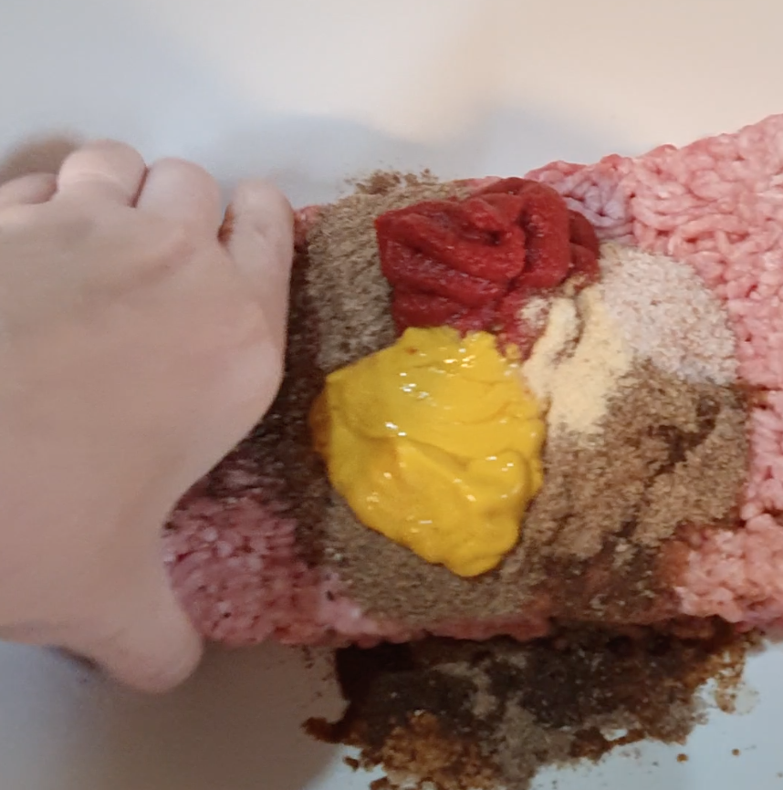 A hand mixes ground meat with mustard, ketchup, more ingredients, and seasonings in a bowl.