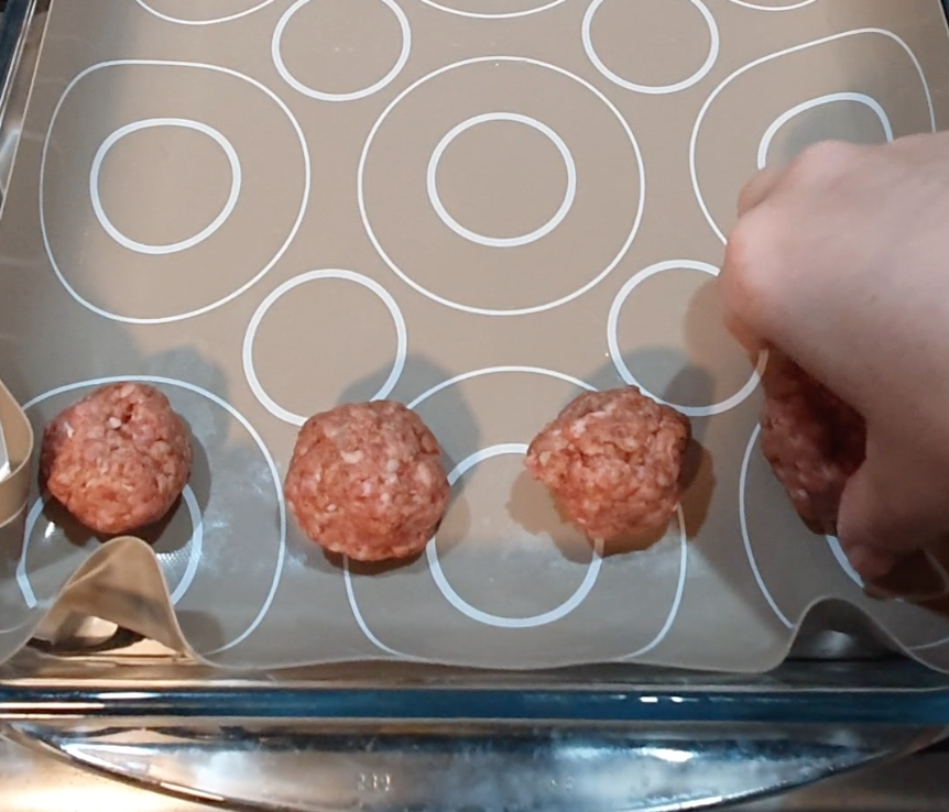 A person's hand placing raw meatballs on a tray.