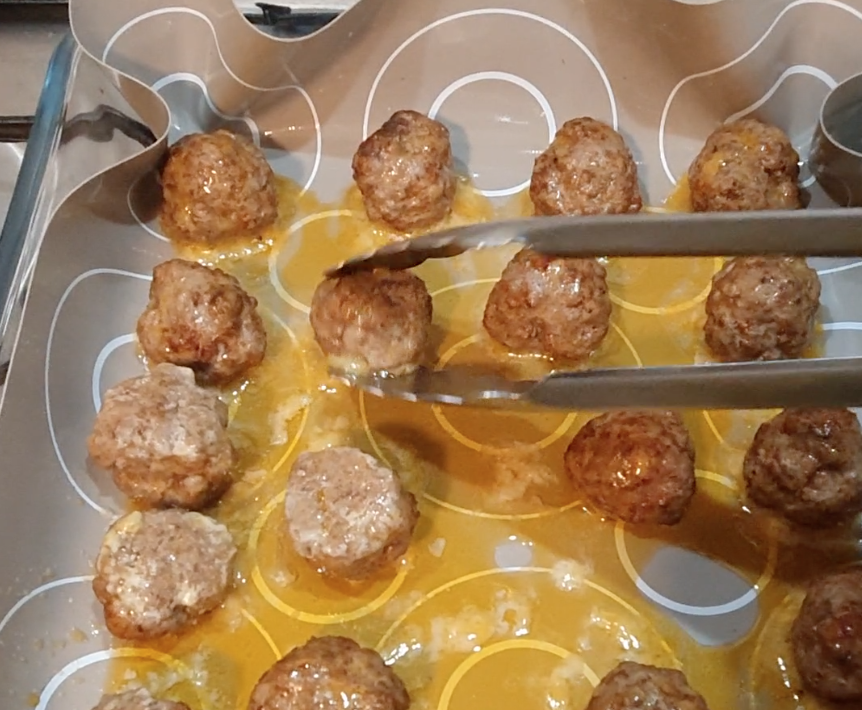 A pair of tongs is picking up a cooked meatball from a baking tray lined with patterned parchment paper.
