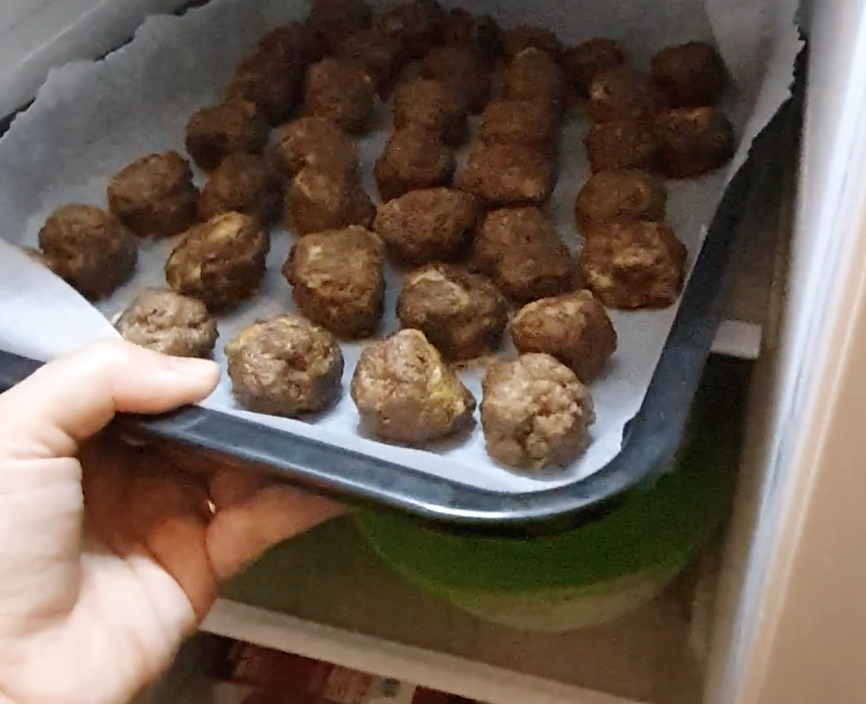 A hand places a baking tray lined with parchment paper and filled with rows of cooked meatballs into a freezer.