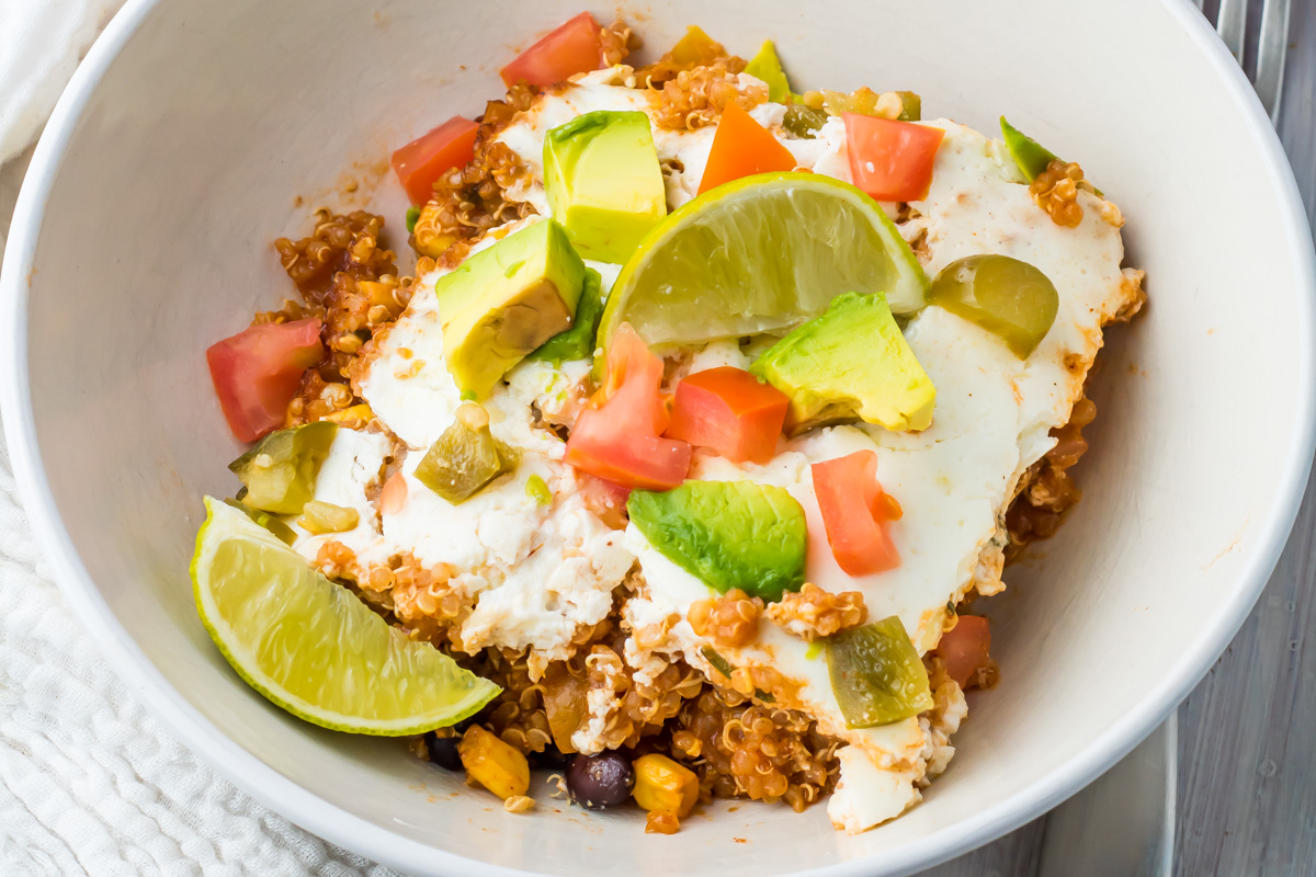 A bowl of quinoa topped with diced tomatoes, avocado, lime wedges, and cottage cheese makes for one of the best high protein recipes on a white surface.