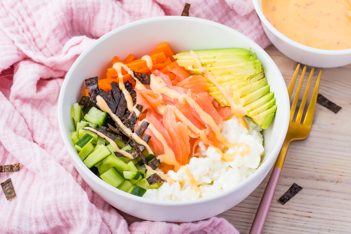 A white bowl contains sliced avocado, tomato, cucumber, carrot, seaweed strips, and rice, drizzled with sauce, on a pink cloth with a fork and dipping sauce nearby.