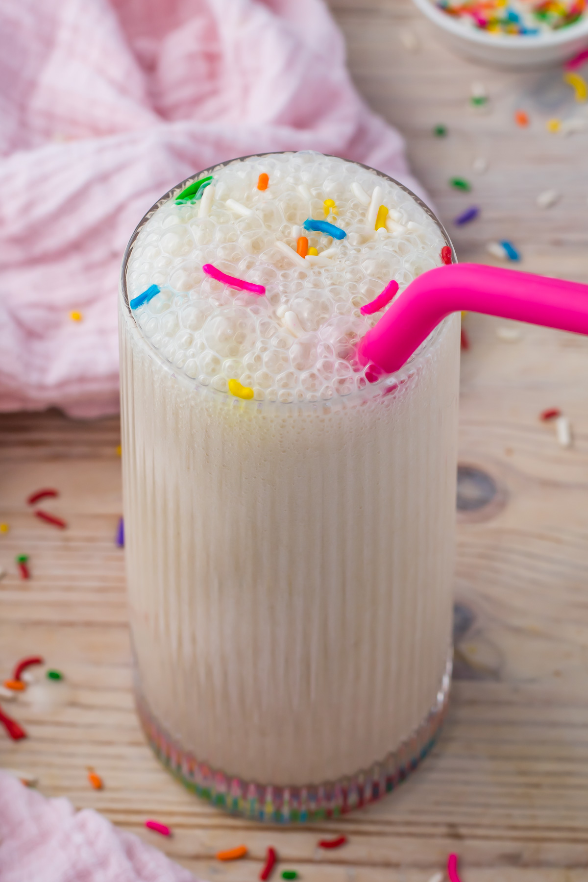 A glass of frothy Cake Batter Protein Shake topped with colorful sprinkles and a pink straw, placed on a wooden surface with more sprinkles scattered around.