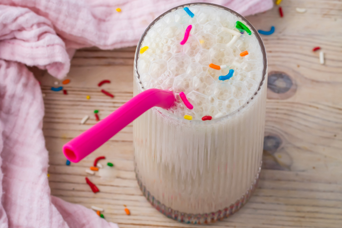 A glass of frothy Cake Batter Protein Shake with colorful sprinkles on top, a pink straw, and a pink cloth on a wooden surface.