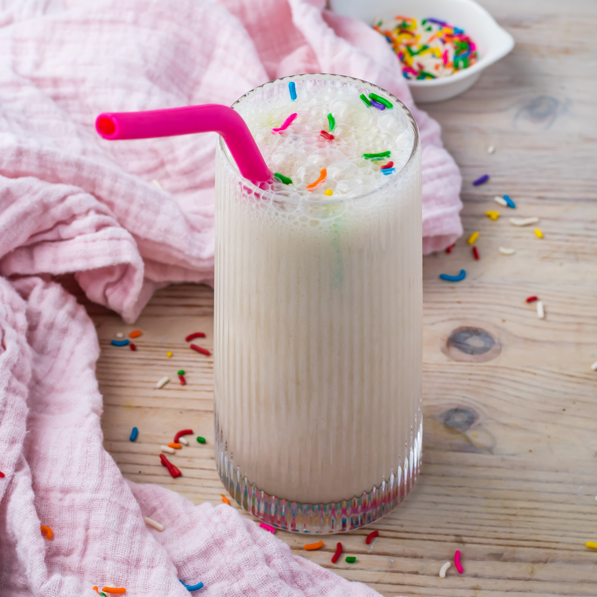 A glass of Cake Batter Protein Shake with a pink straw and colorful sprinkles on top, placed on a wooden table with a pink cloth and scattered sprinkles.