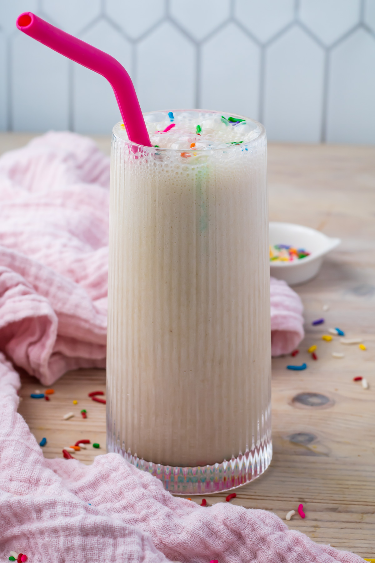 A tall glass of cake batter protein shake with a pink straw, topped with colorful sprinkles, sits on a wooden surface with a pink cloth and a small bowl of sprinkles in the background.