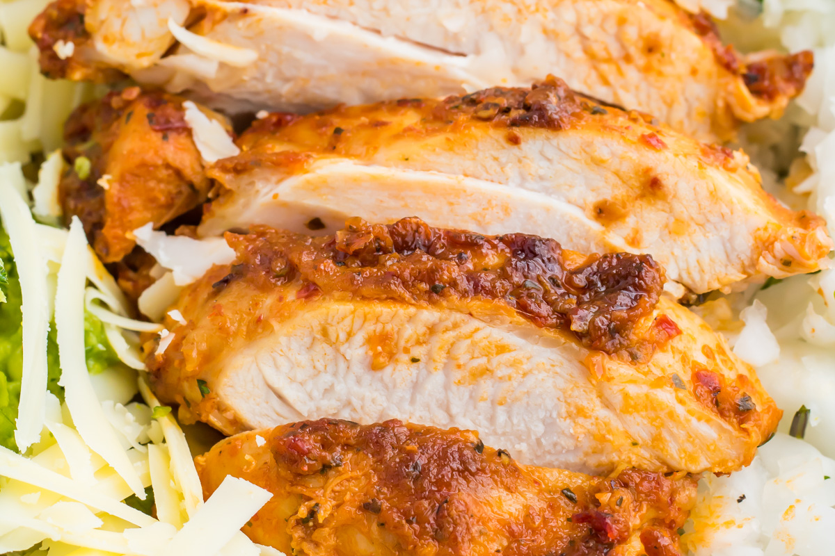 Close-up of sliced, cooked chipotle pollo asado with a seasoned, roasted crust, served on a bed of shredded cheese and vegetables.